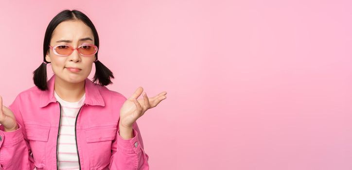 Close up portrait of asian girl looking confused, shrugging puzzled and looking at camera, wearing sunglasses, standing over pink background.