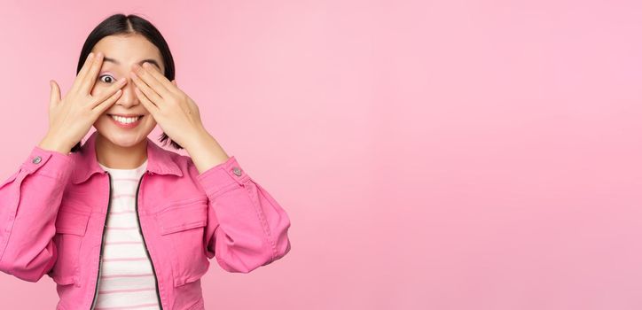Portrait of asian girl peeks with excitement through fingers, covers eyes, seeing surprise, standing over pink background. Copy space
