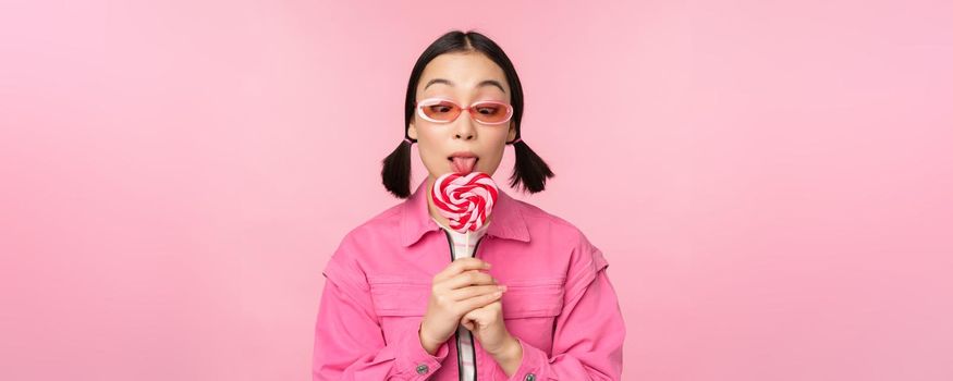 Silly and cute asian female model licking lolipop, eating candy sweet and smiling, looking excited, standing over pink background.