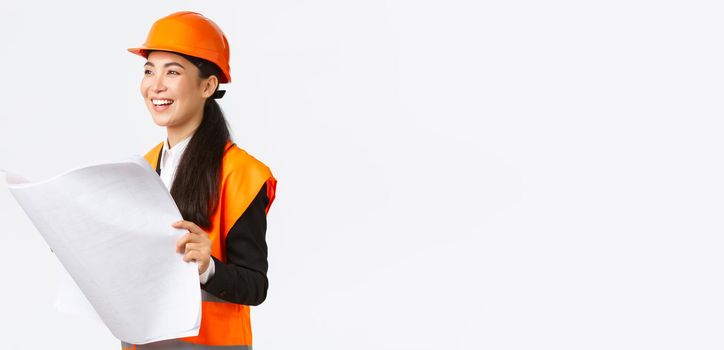 Satisfied happy asian female chief engineer looking at results of construction works, studying blueprint with pleased face, wearing safety helmet and reflective clothings at building zone.