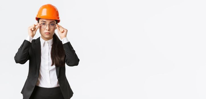Lets get to business. Serious-looking confident female asian chief engineer inspect enterprise, wearing safety helmet and suit, put on glasses, standing white background, ready to work.