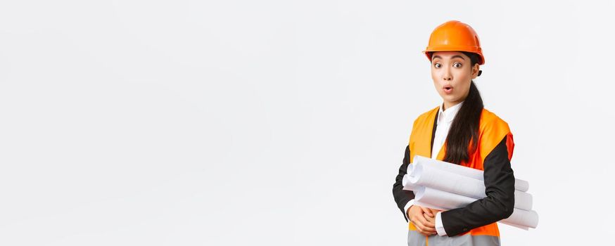 Surprised and excited asian female chief architect in safety helmet and reflective jacket, visit construction area with blueprint, looking amused, listening to interesting suggestion of manager.