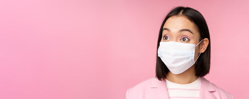 Close-up portrait of asian businesswoman in medical face mask, looking surprised, standing in suit over pink background. Copy space