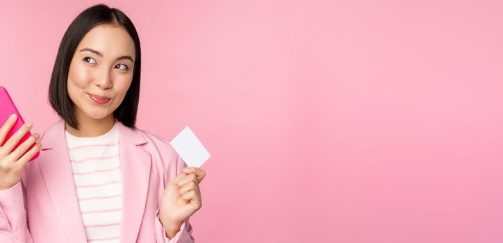 Smiling asian corporate woman, lady in suit thinking, holding smartphone and credit card, plan to buy smth online, shopping with mobile phone, pink background.