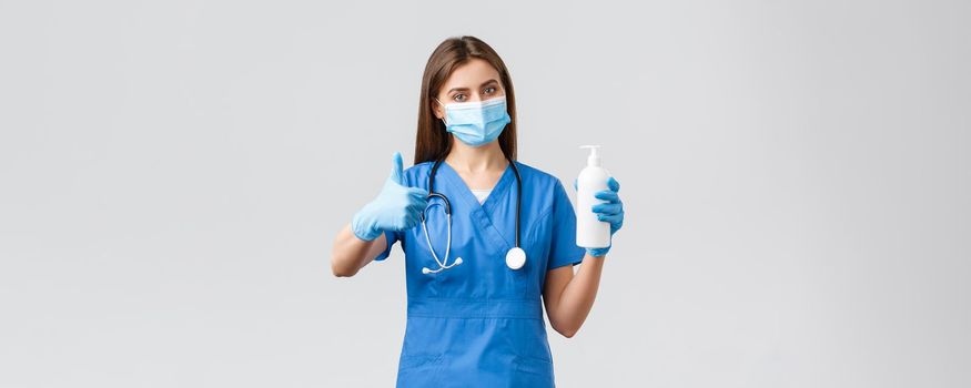 Covid-19, preventing virus, health, healthcare workers and quarantine concept. Confident female doctor, nurse in blue scrubs in medical mask and scrubs advice using hand sanitizer, thumbs-up approval.