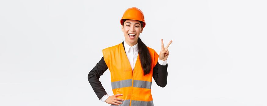 Happy upbeat asian female asian architect in safety helmet and reflective jacket, showing peace sign and smiling, assured in victory of her company, winning tender on construction works.