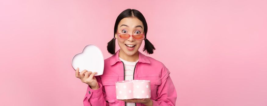 Cute asian girl giving you gift in heart shaped box, kissing and smiling, concept of holiday and celebration, standing over pink background.
