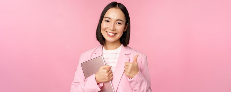 Portrait of corporate woman, girl in office in business suit, holding digital tablet, showing thumbs up, recommending company, standing over pink background.