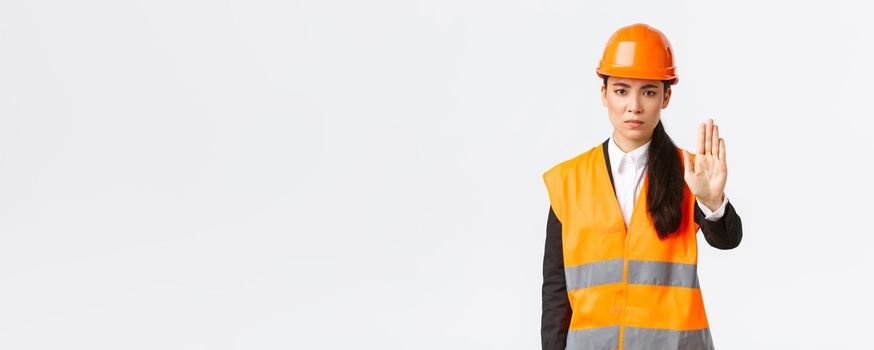 Serious-looking disappointed asian female architect, construction manager at working area wearing safety helmet, showing stop gesture, prohibit action, forbid trespassing, white background.