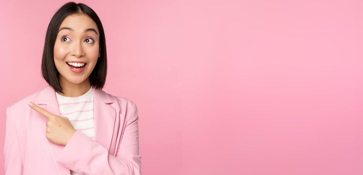 Enthusiastic young asian businesswoman, office employee pointing finger left, looking at banner, advertisement with happy smile, showing advertisement, pink background.