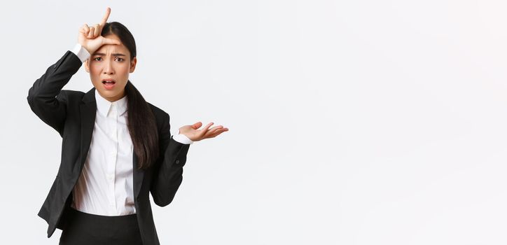 Bothered and annoyed asian businesswoman scolding employee for failure, showing loser gesture and raising hand in dismay, cant understand why so stupid, complaining over white background.