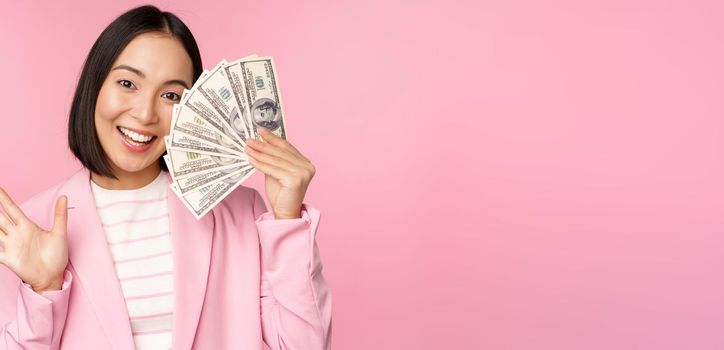 Microcredit, investment and business people concept. Young asian businesswoman, corporate lady showing money, cash dollars, waving hand, pink background.