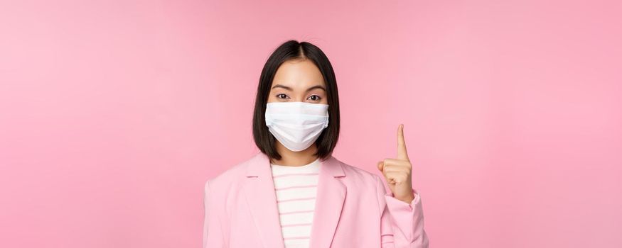 Close up portrait of asian businesswoman in medical face mask and suit, pointing finger up, showing advertisement, top banner, standing over pink background.