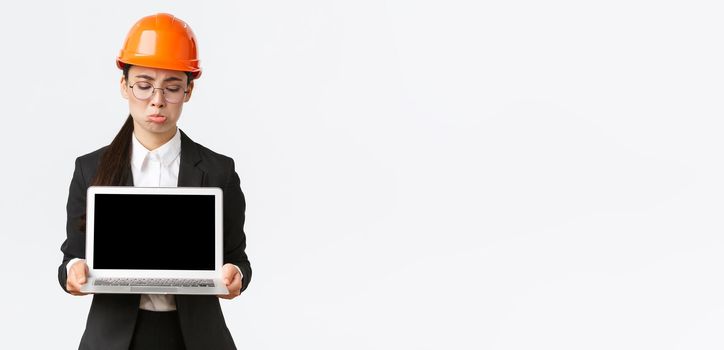 Disappointed gloomy asian female engineer showing bad results, wearing safety helmet and business suit, holding laptop computer and looking at screen displeased, standing white background pouting.