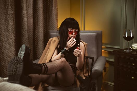 the female criminal in a hotel sits on a chair smoking a cigarette and drinking wine