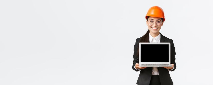 Smiling professional female asian engineer introduce construction plan to investors or clients, standing in safety helmet and suit showing laptop screen with pleased smile, white background.