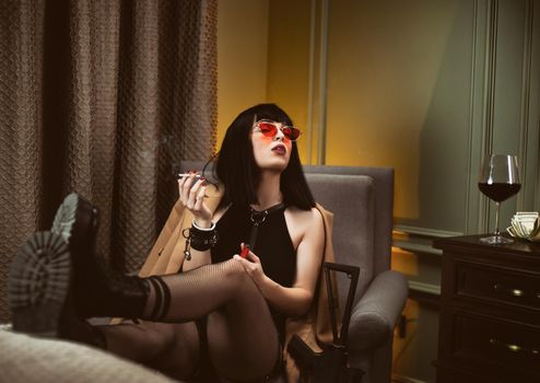 the female criminal in a hotel sits on a chair smoking a cigarette and drinking wine