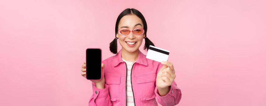 Image of smiling korean woman showing credit card and mobile phone screen, smartphone application interface, paying online, shopping contactless, standing over pink background.