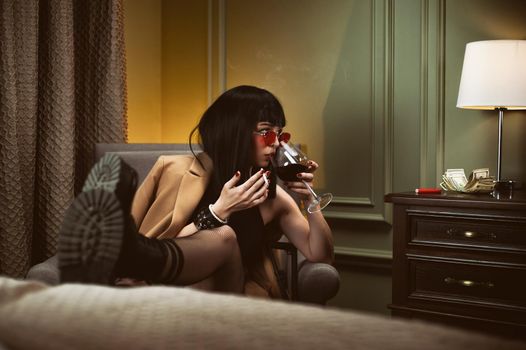 the female criminal in a hotel sits on a chair smoking a cigarette and drinking wine