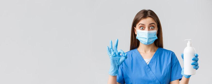 Covid-19, preventing virus, healthcare workers and quarantine concept. Excited and enthusiastic female nurse or doctor in blue scrubs and medical mask recommend soap or hand sanitizer, show okay.