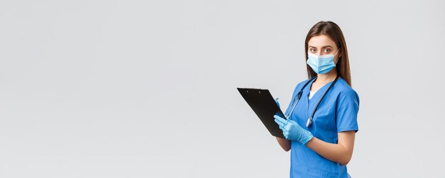 Covid-19, preventing virus, health, healthcare workers and quarantine concept. Professional female nurse or doctor in blue scrubs, medical mask and gloves, writing down patient info using clipboard.