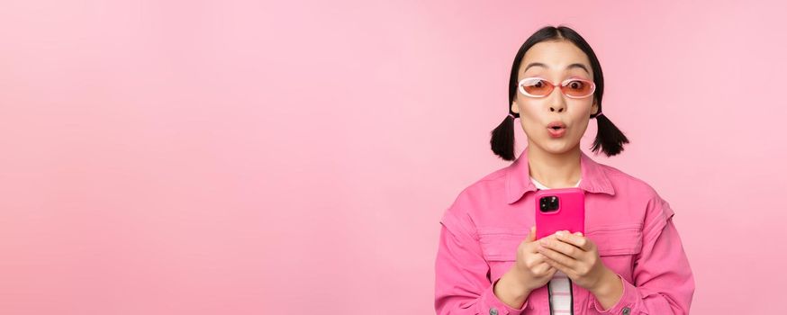 Portrait of korean girl in stylish sunglasses holding mobile phone, using smartphone app, standing over pink background. Copy space