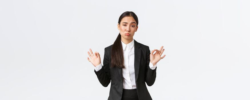 Pleased sassy asian businesswoman in black suit show not bad gesture, nod in approval and make okay gesture, very good work, praising coworker, standing satisfied over white background.