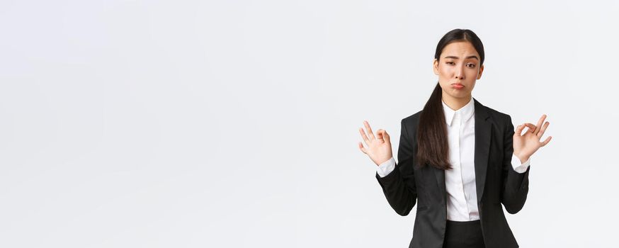 Pleased sassy asian businesswoman in black suit show not bad gesture, nod in approval and make okay gesture, very good work, praising coworker, standing satisfied over white background.