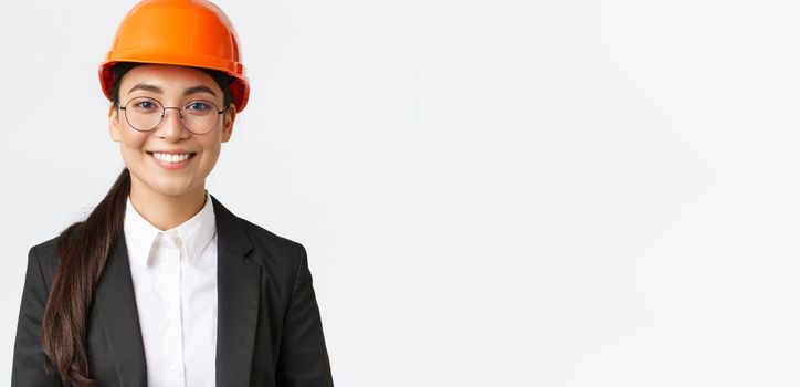 Close-up of smiling professional asian female engineer, businesswoman in suit and safety helmet having inspection at factory, looking confident and pleased, standing white background.