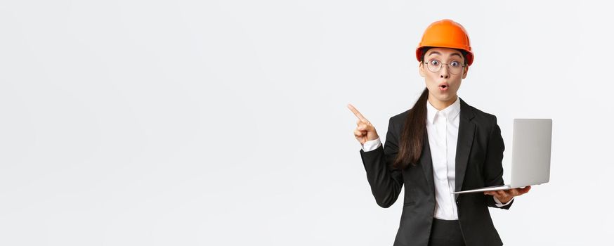 Impressed asian female entrepreneur at factory wearing safety helmet and business suit, pointing finger left and holding laptop computer, showing diagram at enterprise, white background.