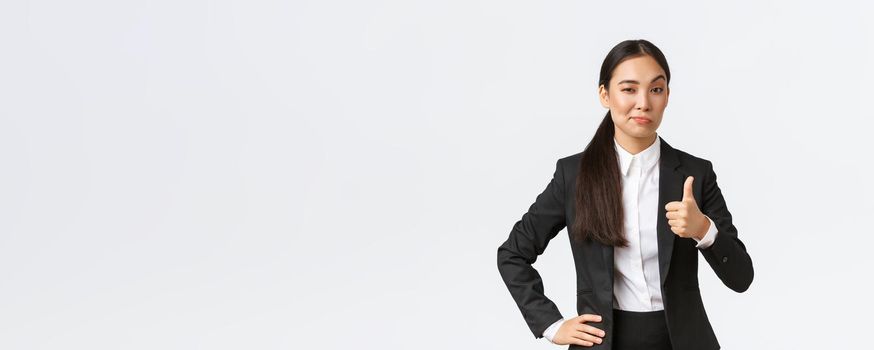 Pleased female entrepreneur, lady boss in black suit satisfied with your work, showing thumbs-up and nod in approval, praise great job, well done, standing white background.