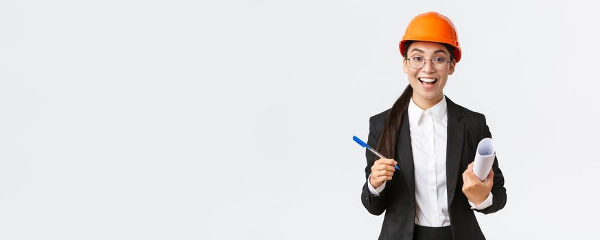 Smiling professional female architect, asian engineer in helmet and business suit, holding blueprints and pen, siging contract, making deal with investors at construction area, white background.