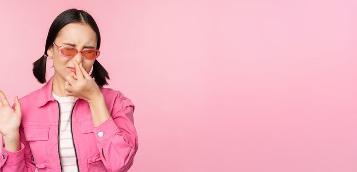 Stinky. Asian girl in sunglasses and stylish outfit, shuts her nose from disgust, dislike bad smell, standing over pink background.