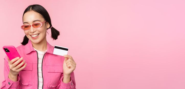 Online shopping. Smiling asian girl shopper, holding smartphone and credit card, paying in mobile app, standing over pink background.