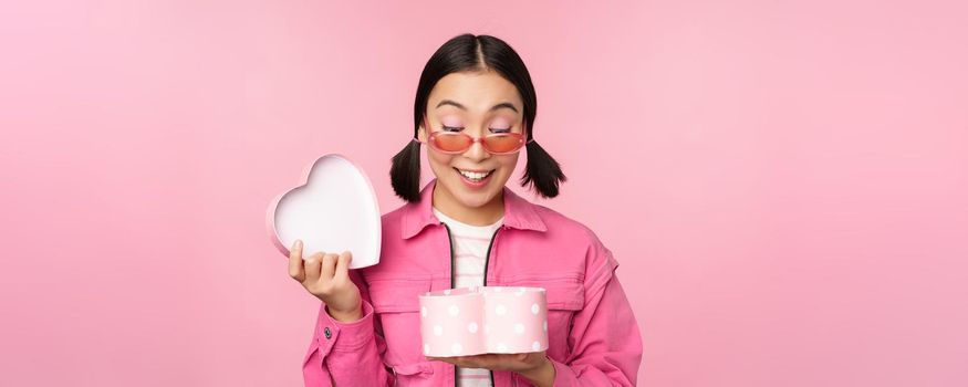 Beautiful asian girl opens up heart shaped gift box, looking happy, standing in sunglasses, smiling surprised at camera, pink background.