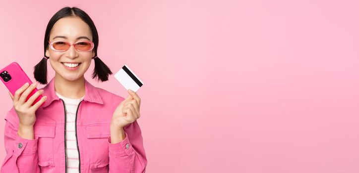 Beautiful korean woman holding smartphone, credit card, smiling at camera, buying online, shopping with mobile phone, standing over pink background.