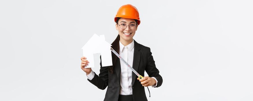 Smiling professional female asian engineer, architect in helmet and business suit showing house maket and tape measure, ready starting home renovation, construction works, white background.