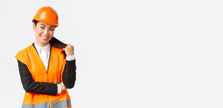 Portrait of confident smiling asian female construction manager, engineer at building area, wearing safety helmet and reflective jacket looking delighted at project, being pleased with finished work.