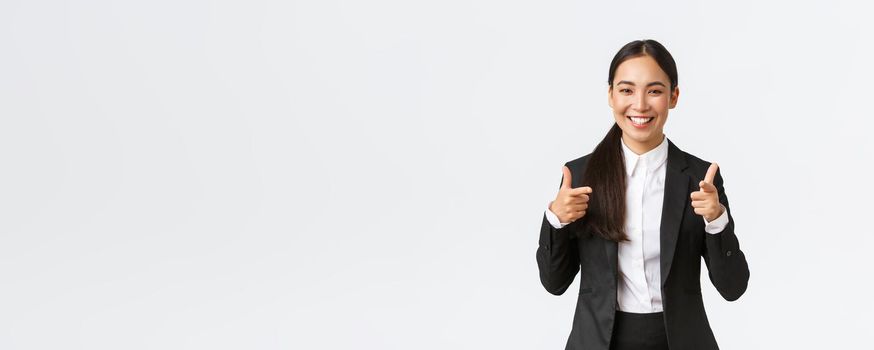Confident smiling asian saleswoman in black suit showing thumbs-up, guarantee quality of product or providing best service. Sales manager trying sell something to client, white background.