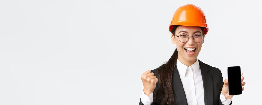 Successful winning asian female engineer, architect in safety helmet and suit, showing smartphone screen, fist pump and shouting yes from rejoice, triumphing over achievement, white background.