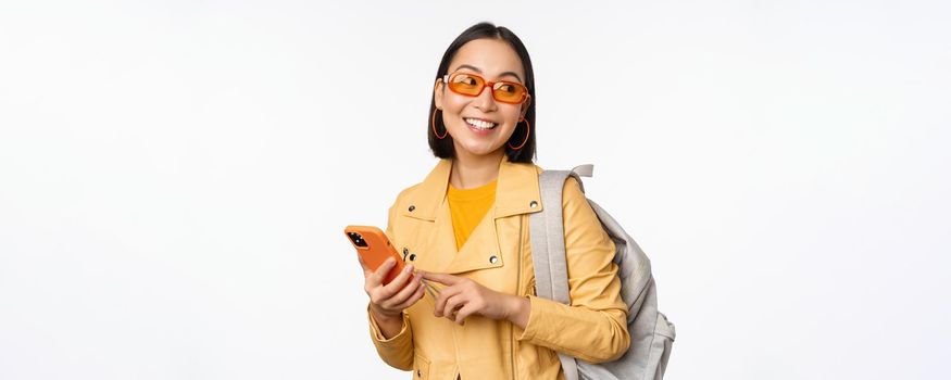 Stylish young asian woman tourist, traveller with backpack and smartphone smiling at camera, posing against white background. Copy space