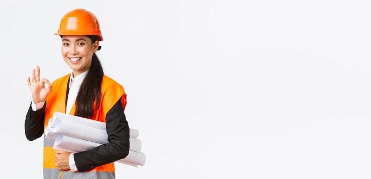 Confident successful female asian construction manager, architect in safety helmet and jacket, showing okay gesture and carry blueprints of building project, guarantee quality, white background.