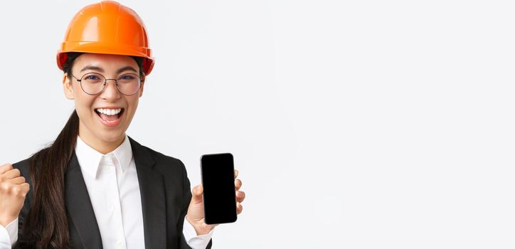 Successful winning asian female engineer, architect in safety helmet and suit, showing smartphone screen, fist pump and shouting yes from rejoice, triumphing over achievement, white background.