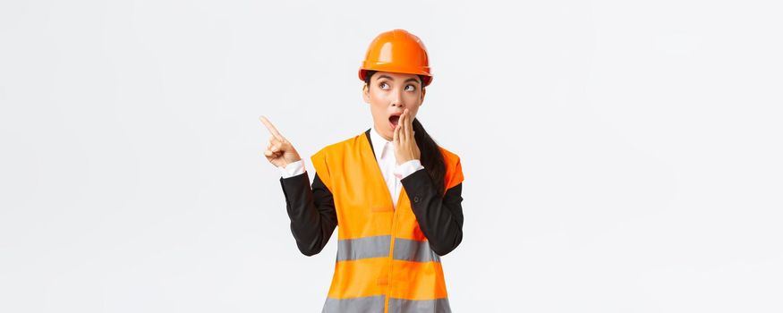 Shocked and impressed asian female engineer, construction manager in safety helmet and reflective jacket, gasping pointing finger upper left corner amazed, found out something interesting.