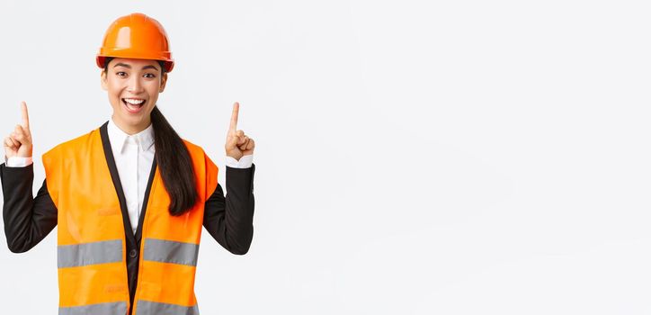 Building, construction and industrial concept. Happy smiling female asian engineer in safety helmet and reflective clothing, introduce new object, estate for sale. Architect pointing fingers up.