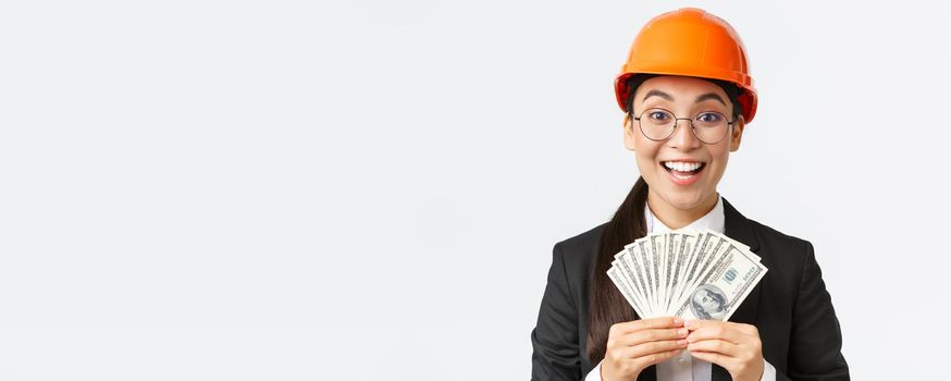 Close-up of happy and excited female asian entrepreneur at factory, wear safety helmet and business suit, showing big income, holding money and rejoicing, smiling pleased, white background.