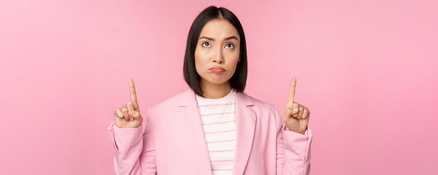 Upset asian corporate woman in office suit, pointing and looking up with disappointed face expression, standing over pink background.