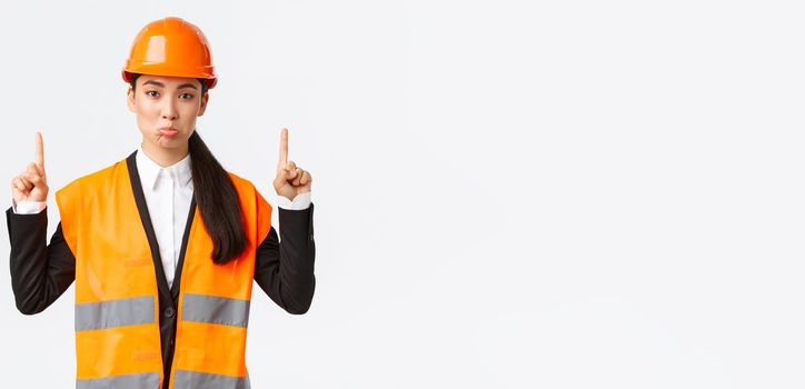 Building, construction and industrial concept. Displeased sad asian female architect complaining, wearing safety helmet and reflective clothing, pouting upset as pointing fingers up.