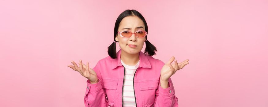 Close up portrait of asian girl looking confused, shrugging puzzled and looking at camera, wearing sunglasses, standing over pink background.