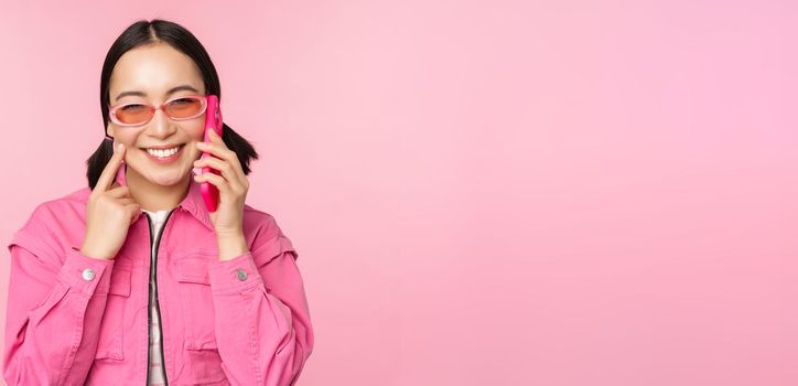 Beautiful korean female model in sunglasses, talking on mobile phone with happy face, using cellular service to call friend on smartphone, standing over pink background.